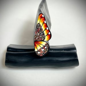 May include: A close-up of a polymer clay sculpture of a butterfly with orange, yellow, white, and brown details. The butterfly is displayed on a black surface.
