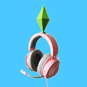 Puede incluir: Auriculares de juego rosas con una parte superior en forma de diamante verde. Los auriculares tienen un micrófono y almohadillas para los oídos con una malla gris.