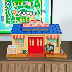 Może przedstawiać: Niebiesko-żółty model zabawki Nook's Cranny z serii gier wideo Animal Crossing. Budynek ma czerwone drzwi, znak z napisem "Nook's Cranny" i dwa okna z białymi markizami.