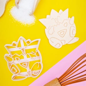 Op de afbeelding: Twee lichtbeige plastic koekjesvormpjes in de vorm van een Togepi-figuur, de ene toont de omtrek en de andere de interne reliëfdetails, geplaatst op een felgeel oppervlak met een houten garde en witte suiker.
