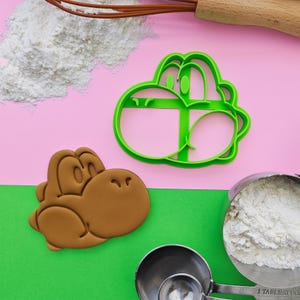 Puede incluir: Cortador de galletas de plástico verde en forma de dinosaurio de dibujos animados con una galleta marrón hecha con el cortador. El cortador de galletas y la galleta están sobre una superficie rosa y verde con harina blanca en el fondo.