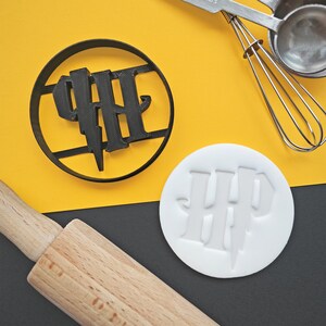 Puede incluir: Un cortador de galletas de plástico negro con las letras "HP" y un rayo. Un sello de fondant blanco con el mismo diseño está al lado. Un rodillo de madera, un batidor y cucharas medidoras también están en la imagen.
