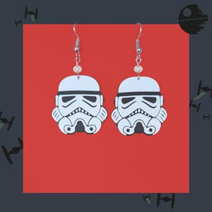 May include: A pair of white and black Stormtrooper earrings. Each earring features a Stormtrooper helmet design with a silver hook and a small, clear bead. The earrings are set against a red background with a dark gray border.