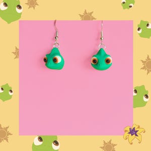 Op de afbeelding: Een paar groene kameleonoorbellen met oranje en witte ogen, hangend aan zilveren haken. De oorbellen zijn geplaatst tegen een roze achtergrond, met een geel en roze patroonrand met zon- en kameleonillustraties.