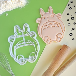 Pode incluir: Dois cortadores de biscoitos em forma de personagem de desenho animado, um branco e outro pêssego, estão em uma superfície verde e amarela. Um batedor e colheres de pau também são visíveis. Farinha polvilhada no canto superior esquerdo.