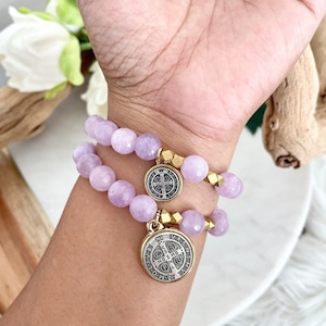 May include: Two lavender beaded bracelets with gold accents and Saint Benedict medals. The bracelets are made of round, faceted beads and are worn on a wrist. The medals are circular with detailed engravings.