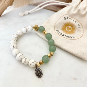 May include: A white and green beaded bracelet with a gold cross charm and a gold charm with a religious image. The bracelet is on a white surface and comes with a small white drawstring bag with the text "Give Love Blessings".