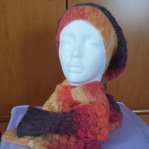 Op de afbeelding: Een gehaakte set van muts en sjaal in tinten oranje, geel en bruin. De muts is van het type baret en de sjaal is een lang, vloeiend ontwerp.