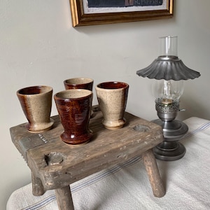 May include: A set of four ceramic cups with a speckled beige and dark brown glazed finish, arranged on a weathered wooden stool. The cups feature a flared rim and a tapered base, showcasing a two-tone color scheme with a smooth, glossy texture.