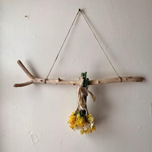 May include: A rustic wall hanging featuring a natural driftwood branch suspended by twine. A bundle of yellow and white flowers is tied to the branch with twine and green stems. The wall is painted a light beige color.
