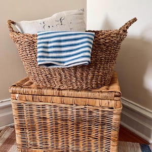 Peut inclure: Deux paniers en osier tress&eacute;, l'un ovale avec des poign&eacute;es et l'autre rectangulaire avec un couvercle &agrave; charni&egrave;re. Un tissu ray&eacute; bleu et blanc et un oreiller en lin avec du texte d&eacute;lav&eacute; reposent dans le panier sup&eacute;rieur. Les paniers sont pos&eacute;s sur un tapis ray&eacute;.