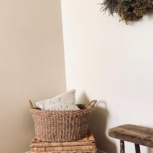 Peut inclure: Un panier en osier tress&eacute; avec des poign&eacute;es est pos&eacute; sur un coffre en osier assorti. Un coussin d&eacute;coratif repose &agrave; l'int&eacute;rieur du panier. Un petit banc en bois et des fleurs s&eacute;ch&eacute;es sont &eacute;galement visibles.