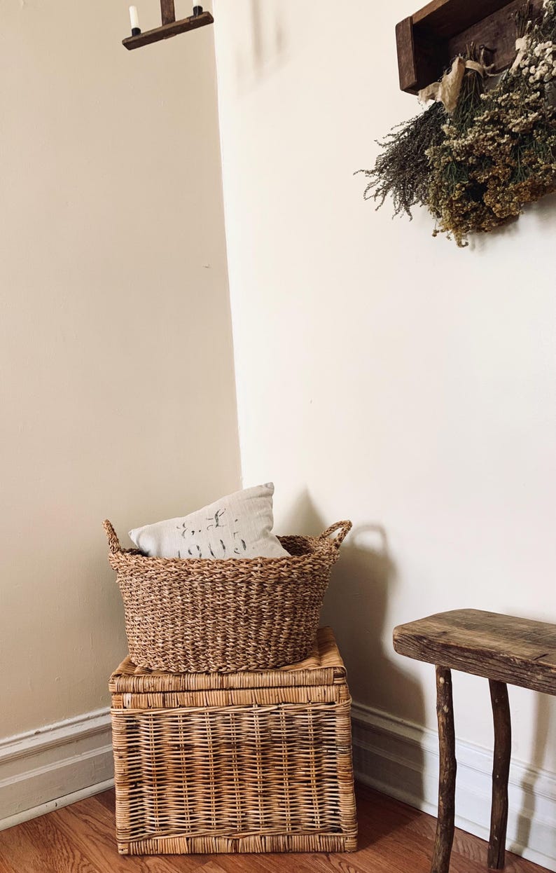 Peut inclure: Un panier en osier tress&eacute; avec des poign&eacute;es est pos&eacute; sur un coffre en osier assorti. Un coussin d&eacute;coratif repose &agrave; l'int&eacute;rieur du panier. Un petit banc en bois et des fleurs s&eacute;ch&eacute;es sont &eacute;galement visibles.
