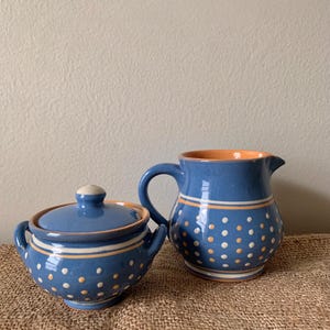 May include: Blue ceramic sugar bowl with lid and a matching creamer pitcher. Both pieces feature a blue glaze with white and orange polka dots and stripes. The sugar bowl has two handles and the creamer has a handle and spout.