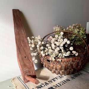 May include: A rustic, wooden butter churn paddle with a handle and a peg. A woven basket filled with dried flowers sits nearby on a burlap sack with text. The scene evokes a vintage, farmhouse aesthetic.