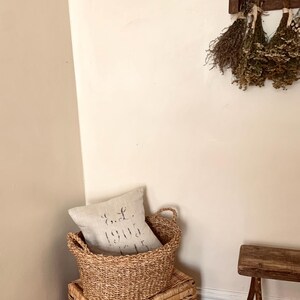 Peut inclure: Un panier et un coffre de rangement en osier sont empil&eacute;s, avec un oreiller en lin &agrave; l'int&eacute;rieur du panier. L'oreiller porte le texte "E.L. 1905". La sc&egrave;ne se d&eacute;roule dans un coin avec un banc en bois et des herbes s&eacute;ch&eacute;es accroch&eacute;es au mur.