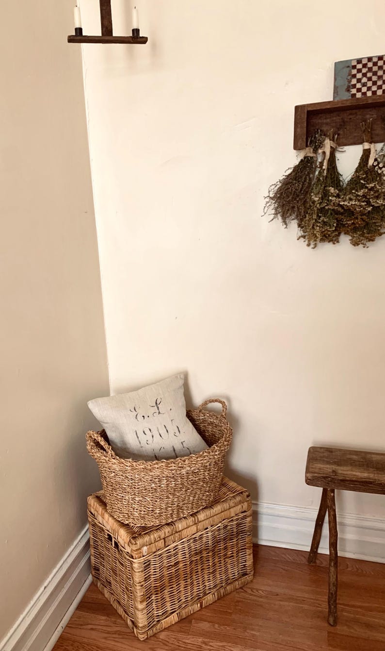 Peut inclure: Un panier et un coffre de rangement en osier sont empil&eacute;s, avec un oreiller en lin &agrave; l'int&eacute;rieur du panier. L'oreiller porte le texte "E.L. 1905". La sc&egrave;ne se d&eacute;roule dans un coin avec un banc en bois et des herbes s&eacute;ch&eacute;es accroch&eacute;es au mur.