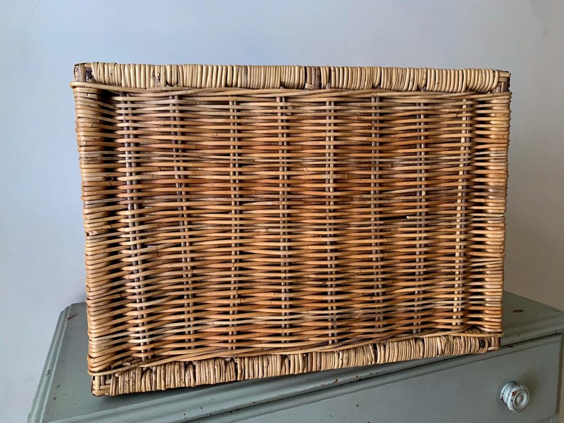Peut inclure: Un panier rectangulaire en osier de couleur marron clair naturel. Le panier est tiss&eacute; de mani&egrave;re serr&eacute;e avec des brins horizontaux et verticaux, cr&eacute;ant une surface textur&eacute;e. Le panier est pos&eacute; sur une surface bleu clair.