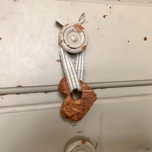 May include: A textured, brown, heart-shaped ornament hangs from a light blue and white striped fabric ribbon. The ribbon is secured to a circular, light gray knob with visible paint chips, which is attached to a light gray drawer front.