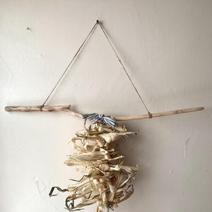 May include: A rustic wall hanging featuring dried corn husks suspended from a driftwood branch. The husks are tied together and accented with a blue and white striped ribbon. The piece is hung by twine from a nail in the wall, creating a natural, earthy aesthetic.