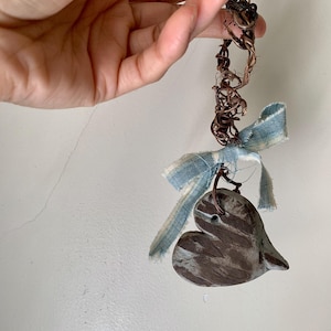 May include: A heart-shaped ornament made of dark, textured material with a hole at the top. It is suspended by a twisted brown vine-like cord and decorated with a frayed, light blue and white striped fabric ribbon tied in a bow.