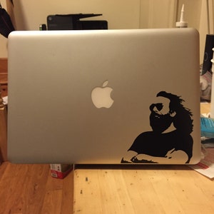 May include: A silver laptop computer with a black and white sticker of a man with a beard and long hair. The sticker is on the right side of the laptop. The Apple logo is on the left side of the laptop.