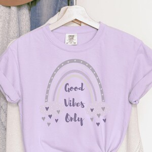 May include: A light purple t-shirt with rolled-up sleeves. The shirt features a rainbow design with the words "Good Vibes Only" in a script font. Small heart shapes are below the rainbow.