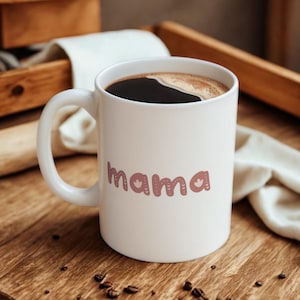 May include: A white ceramic mug filled with dark coffee. The word "mama" is printed on the front in a decorative, mauve-colored font with floral accents. The mug sits on a wooden surface.