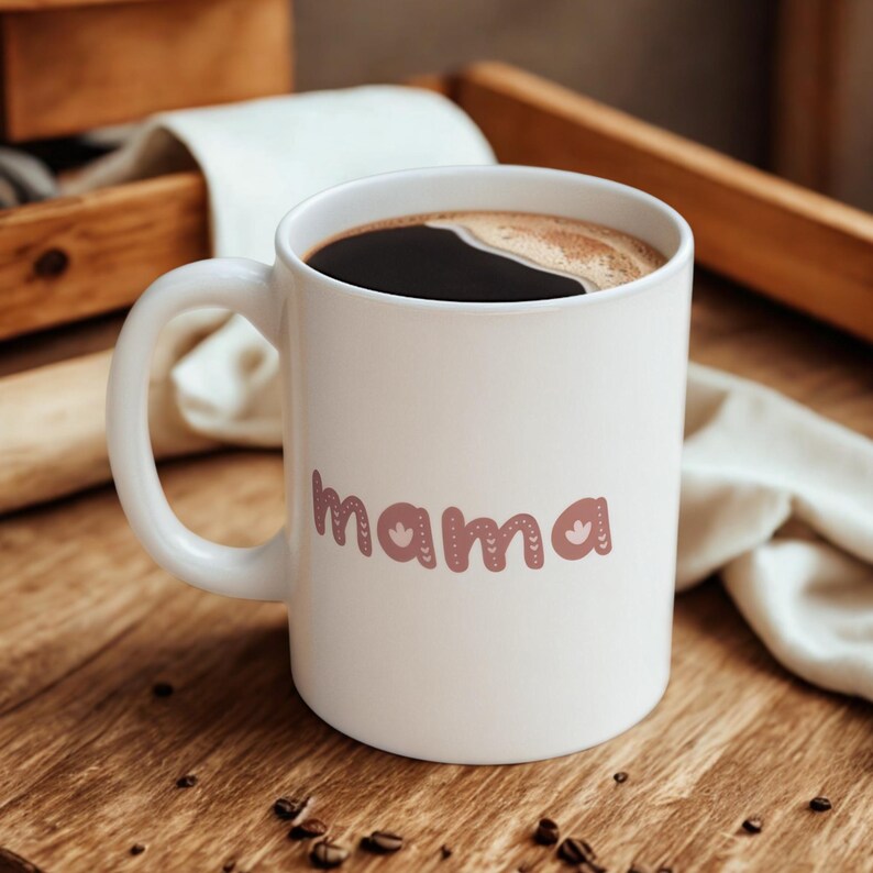 May include: A white ceramic mug filled with dark coffee. The word "mama" is printed on the front in a decorative, mauve-colored font with floral accents. The mug sits on a wooden surface.