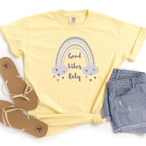 May include: A light yellow t-shirt with a rainbow graphic and the text "Good Vibes Only". The rainbow is light blue and purple, with clouds and hearts. The shirt is paired with denim shorts and brown sandals.
