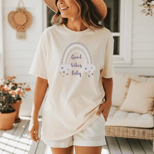 May include: Cream-colored t-shirt with a rainbow graphic and the text "Good Vibes Only." The rainbow design features shades of gray, purple, and white. The shirt is paired with white shorts. The overall aesthetic is casual and relaxed.