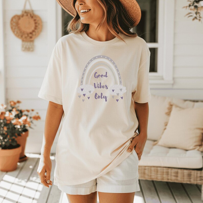 May include: Cream-colored t-shirt with a rainbow graphic and the text "Good Vibes Only." The rainbow design features shades of gray, purple, and white. The shirt is paired with white shorts. The overall aesthetic is casual and relaxed.