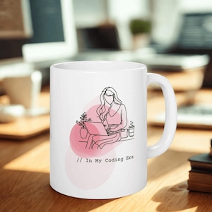 May include: White ceramic mug featuring a line art illustration of a person working on a laptop, with a pink background. The text "// In My Coding Era" is printed below the illustration. A coffee cup and plant are also depicted.