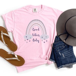 May include: A light pink t-shirt with a rainbow graphic and the words "Good Vibes Only." The rainbow is gray, lavender, and white. The shirt is paired with denim shorts, a gray hat, and brown sandals.
