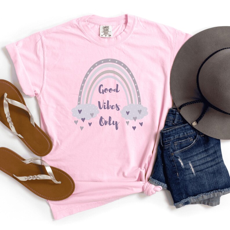 May include: A light pink t-shirt with a rainbow graphic and the words "Good Vibes Only." The rainbow is gray, lavender, and white. The shirt is paired with denim shorts, a gray hat, and brown sandals.