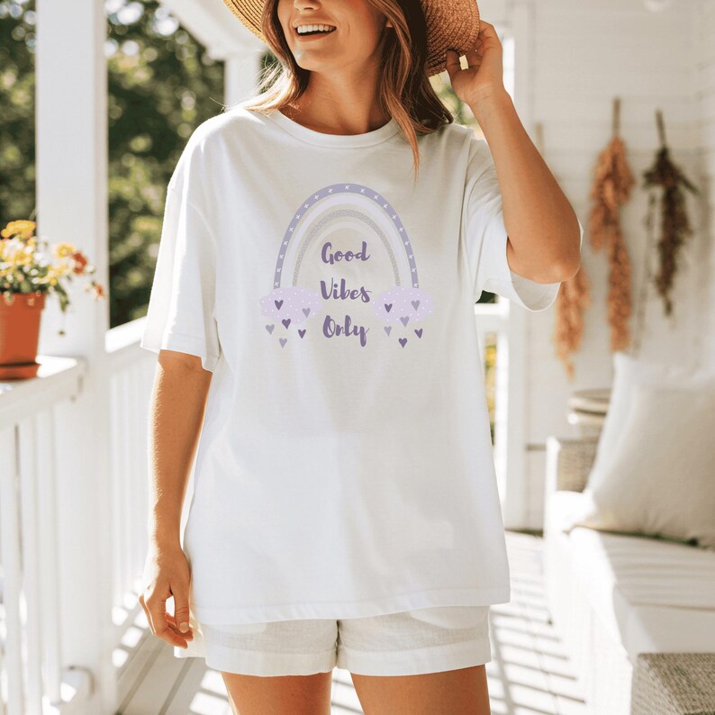 May include: White t-shirt with a pastel rainbow graphic and the text "Good Vibes Only." The design includes purple clouds and hearts. The shirt is a casual, oversized fit.