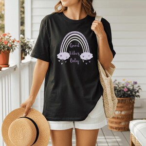 May include: Black t-shirt featuring a rainbow design with the words "Good Vibes Only" in a whimsical font. The rainbow has a purple and white color scheme, with heart accents. The shirt is paired with white shorts and a straw hat.