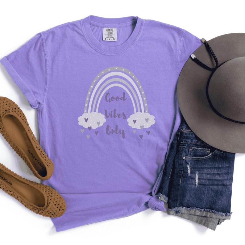 May include: A lilac-colored t-shirt with a rainbow graphic and the text "Good Vibes Only." The rainbow design features silver and gray arches, with fluffy clouds and heart accents. A gray hat and denim shorts are visible.