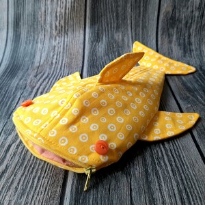 May include: A yellow fabric shark pencil case with white polka dots and orange buttons. The zipper is open and the lining is pink.