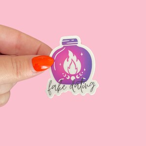 May include: A white sticker with a purple and pink gradient design of a jar with a flame inside. The text "fake dating" is written below the jar.