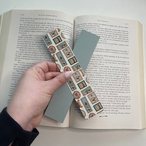 May include: Two bookmarks with a floral and geometric design. The floral bookmark has a pink, green, and white design with a potted plant and books. The geometric bookmark is light blue with a white border.