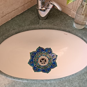 May include: A white oval bathroom sink with a decorative blue and teal floral design around the drain. A chrome faucet is positioned above the sink, and a small glass bottle with a plant is visible in the background.