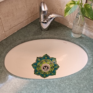 May include: White oval bathroom sink with a decorative floral design in shades of green and teal around the drain. A chrome faucet is mounted on the wall. A small glass bottle with a plant is on the counter.