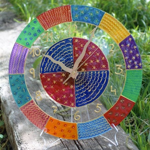 Puede incluir: Un reloj de vidrio colorido con un borde dorado y números dorados. La esfera del reloj está dividida en doce secciones, cada una con un patrón diferente. Las manecillas del reloj son doradas.