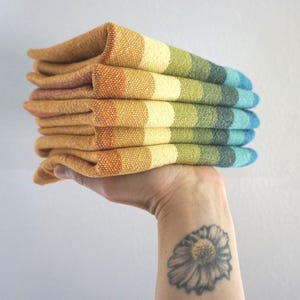May include: A stack of folded textile napkins featuring a woven gradient pattern that transitions through shades of orange, yellow, light green, dark green, and blue. The fabric has a visible textured weave and is stacked in a neat pile.