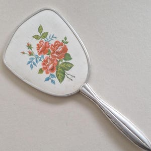 Könnte beinhalten: Ein Vintage-Handspiegel mit silberfarbenem Griff und Rahmen. Das Spiegelgesicht zeigt ein Blumenstickerei-Design mit roten Rosen, grünen Blättern und blauen Akzenten. Ein klassisches Accessoire für den Schminktisch.