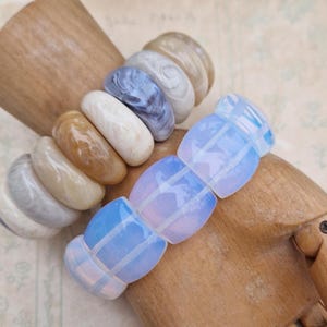 May include: A bracelet with a mix of oval beads in white, beige, and blue hues. The bracelet is displayed on a wooden hand, highlighting the different textures and colours of the beads. The beads are made of various materials.