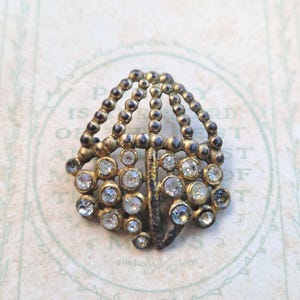 Puede incluir: Broche vintage dorado con diseño en forma de abanico. El broche presenta pequeños brillantes redondos y transparentes engastados en un marco metálico decorativo. La parte superior tiene un diseño de cuentas, lo que aumenta su encanto antiguo.