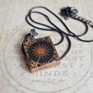 May include: A rectangular pendant necklace with a black chain. The pendant features a detailed, circular design in shades of brown and gold, set against a dark background. The pendant is made of wood and has a silver-colored bail.