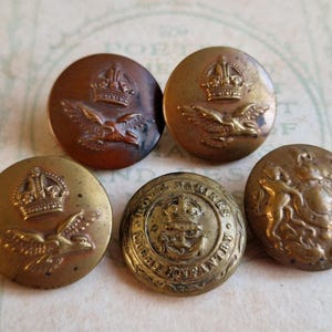 Może przedstawiać: Pięć zabytkowych mosiężnych guzików z szczegółowymi wzorami. Guziki przedstawiają korony, orły i herby. Guziki są w różnych odcieniach złota i brązu, wykazując oznaki wieku i zużycia, sugerując zabytkowe akcesoria wojskowe lub formalne.